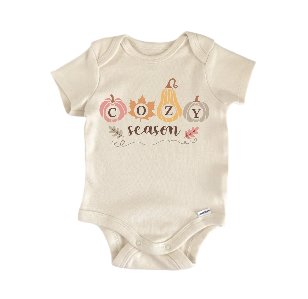 A beige baby onesie with the word "COZY season" printed on it, along with autumn-themed illustrations of pumpkins and leaves.