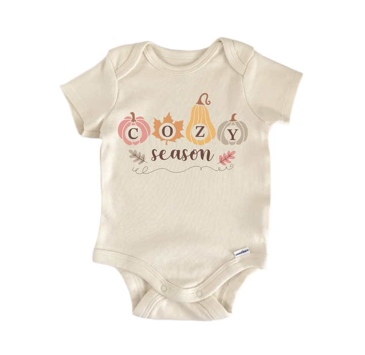 A beige baby onesie with the word "COZY season" printed on it, along with autumn-themed illustrations of pumpkins and leaves.