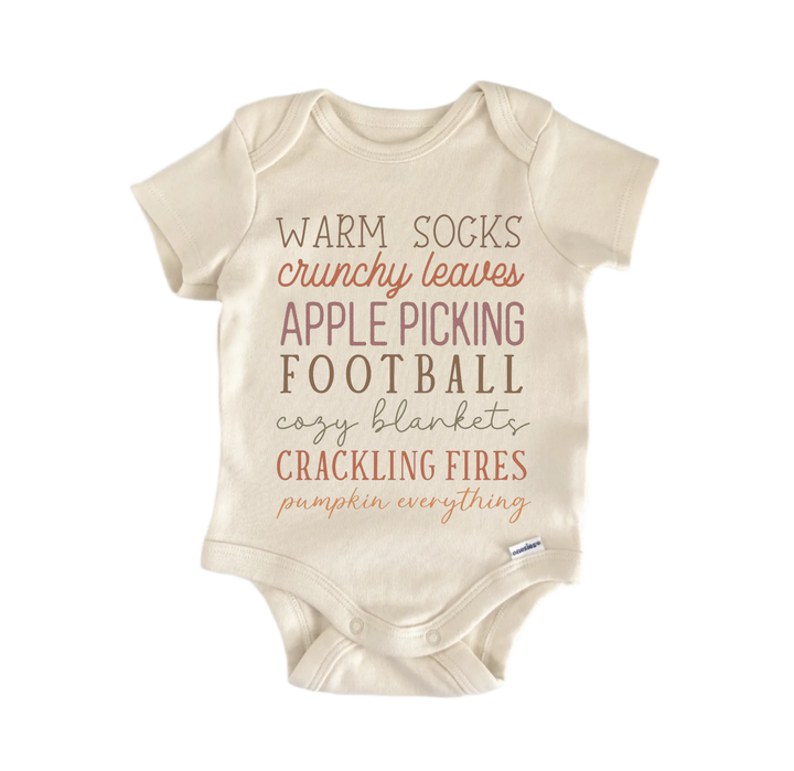 A beige baby onesie with text printed on it, including words like "Warm socks", "crunchy leaves", "apple picking", "football", "cozy blankets", "cracking fires", and "pumpkin everything".